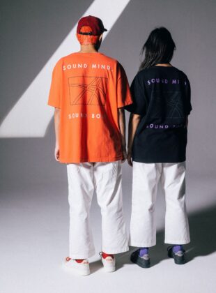 foto de dois modelos de costas, um com camiseta laranja e outra com uma azul-marinho. Ambos vestem calça branca de Kimono. As peças são da parceria entre Zee.Dog Human e Asics.