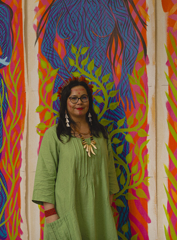 daiara tukano, mulher indígena vestida de verde, em frente a três telas representando mulheres em cores vivas