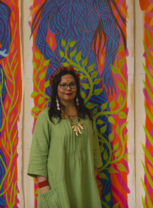 daiara tukano, mulher indígena vestida de verde, em frente a três telas representando mulheres em cores vivas