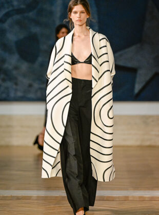 Fendi, inverno 2026 alta-costura Na primeira coleção de alta-costura de Maria Grazia Chiuri como diretora criativa da Fendi, a estilista repete fórmulas de sucesso da própria carreira e revisita os arquivos da grife italiana. A apresentação de inverno 2026 de alta-costura da Fendi foi realizada nesta quinta-feira (09.07) na Galleria Nazionale d'Arte Moderna e Contemporanea (GNAMC), em Roma. O motivo da locação é a reativação de uma exposição feita pela grife ali mesmo, em 1985. Na época, Karl Lagerfeld comemorava duas décadas de parceria com as irmãs Paola, Anna, Franca, Carla e Alda Fendi, então proprietárias e diretoras executivas da empresa. Leia mais: Fendi, inverno 2026 O evento também marcou o retorno da Fendi com um desfile da linha couture, o que não acontecia desde 2024, quando Kim Jones era diretor criativo. E Maria Grazia Chiuri, que assumiu tal posição na casa no final do ano passado, revelou pela primeira vez propostas exclusivas e sob medida na maison. Foi a própria designer quem deu a ideia de reabrir a mostra. Chamada de After Un Percorso di Lavoro. Fendi/Karl Lagerfeld 1985. After Steps Through Work, a exibição fica aberta ao público entre o dia 10 de julho e 25 de outubro de 2026. Quem estiver na capital italiana durante o período poderá conferir criações históricas, croquis e outros documentos. Os itens ajudam a entender o legado que Maria Grazia recupera na passarela. A designer olhou, por exemplo, para um casaco branco com linhas negras que formam um labirinto por toda a superfície – e ele foi recriado no look 08. O mesmo item vintage impulsiona uma pesquisa atual sobre a relação da Fendi com a geometria. A conexão gráfica deriva do interesse de Karl pela Secessão Vienense, movimento artístico do final do século 19 próximo à estética Art Déco. Caracterizada por formas como quadrados, losangos, triângulos e círculos, a vertente teve como expoentes os artistas Gustav Klimt e Egon Schiele, e o arquiteto Josef Hoffmann. O primeiro look da coleção é baseado em uma foto da austríaca Emilie Flöge usando um vestido amplo e listrado. Musa e companheira de vida de Klimt, ela foi uma estilista do início do século 20. Leia mais: Fendi, verão 2026 Esse visual de abertura é usado pela atriz australiana Leila George, que estrela o curta Love Monster (2026), exibido antes do desfile. O uso de produções cinematográficas era comum na trajetória de Maria Grazia como diretora criativa da Dior (2016-2025), mas a obra também referencia Karl. Trata-se de uma alusão a Histoire d’eau (1977), de Jacques De Bascher, considerado o primeiro fashion film da história, encomendado pelo Kaiser no passado e que integra agora a mostra na GNAMC. A história do curta – que acompanha uma alemã por Roma nos anos 1970 – inspira as silhuetas de quimono das jaquetas e sobretudos, além do uso de peleteria em capas e mantos. Da própria imagem de Karl, vêm ainda as chokers em formato de gola de camisa e a clássica cartela em preto e branco – homenagens que a diretora criativa já havia feito em sua estreia no prêt-à-porter em fevereiro. Leia mais: Fendi, verão 2025 Contudo, a estilista não se restringe ao passado da marca com o estilista. Ela revisita códigos bem-sucedidos de sua trajetória: lingeries aparentes sob robes, slip dresses com hot pants visíveis e uma alfaiataria que transita entre o masculino e o feminino. Os vestidos leves e transparentes, assinatura da italiana, são feitos de renda, tule, georgette, chiffon e misturas de seda e lã. A imagem geral pode não surpreender, mas se conecta ao desejo couture nessa temporada por simplicidade e roupas ligadas à intimidade – noções que Maria Grazia domina há tempos. Leia também: Fendi, de Karl Lagerfeld a Kim Jones