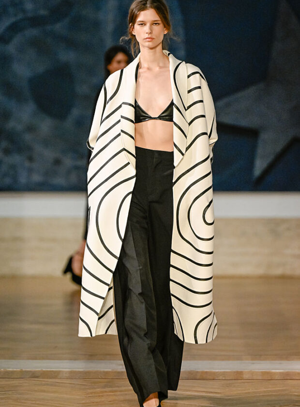 Fendi, inverno 2026 alta-costura Na primeira coleção de alta-costura de Maria Grazia Chiuri como diretora criativa da Fendi, a estilista repete fórmulas de sucesso da própria carreira e revisita os arquivos da grife italiana. A apresentação de inverno 2026 de alta-costura da Fendi foi realizada nesta quinta-feira (09.07) na Galleria Nazionale d'Arte Moderna e Contemporanea (GNAMC), em Roma. O motivo da locação é a reativação de uma exposição feita pela grife ali mesmo, em 1985. Na época, Karl Lagerfeld comemorava duas décadas de parceria com as irmãs Paola, Anna, Franca, Carla e Alda Fendi, então proprietárias e diretoras executivas da empresa. Leia mais: Fendi, inverno 2026 O evento também marcou o retorno da Fendi com um desfile da linha couture, o que não acontecia desde 2024, quando Kim Jones era diretor criativo. E Maria Grazia Chiuri, que assumiu tal posição na casa no final do ano passado, revelou pela primeira vez propostas exclusivas e sob medida na maison. Foi a própria designer quem deu a ideia de reabrir a mostra. Chamada de After Un Percorso di Lavoro. Fendi/Karl Lagerfeld 1985. After Steps Through Work, a exibição fica aberta ao público entre o dia 10 de julho e 25 de outubro de 2026. Quem estiver na capital italiana durante o período poderá conferir criações históricas, croquis e outros documentos. Os itens ajudam a entender o legado que Maria Grazia recupera na passarela. A designer olhou, por exemplo, para um casaco branco com linhas negras que formam um labirinto por toda a superfície – e ele foi recriado no look 08. O mesmo item vintage impulsiona uma pesquisa atual sobre a relação da Fendi com a geometria. A conexão gráfica deriva do interesse de Karl pela Secessão Vienense, movimento artístico do final do século 19 próximo à estética Art Déco. Caracterizada por formas como quadrados, losangos, triângulos e círculos, a vertente teve como expoentes os artistas Gustav Klimt e Egon Schiele, e o arquiteto Josef Hoffmann. O primeiro look da coleção é baseado em uma foto da austríaca Emilie Flöge usando um vestido amplo e listrado. Musa e companheira de vida de Klimt, ela foi uma estilista do início do século 20. Leia mais: Fendi, verão 2026 Esse visual de abertura é usado pela atriz australiana Leila George, que estrela o curta Love Monster (2026), exibido antes do desfile. O uso de produções cinematográficas era comum na trajetória de Maria Grazia como diretora criativa da Dior (2016-2025), mas a obra também referencia Karl. Trata-se de uma alusão a Histoire d’eau (1977), de Jacques De Bascher, considerado o primeiro fashion film da história, encomendado pelo Kaiser no passado e que integra agora a mostra na GNAMC. A história do curta – que acompanha uma alemã por Roma nos anos 1970 – inspira as silhuetas de quimono das jaquetas e sobretudos, além do uso de peleteria em capas e mantos. Da própria imagem de Karl, vêm ainda as chokers em formato de gola de camisa e a clássica cartela em preto e branco – homenagens que a diretora criativa já havia feito em sua estreia no prêt-à-porter em fevereiro. Leia mais: Fendi, verão 2025 Contudo, a estilista não se restringe ao passado da marca com o estilista. Ela revisita códigos bem-sucedidos de sua trajetória: lingeries aparentes sob robes, slip dresses com hot pants visíveis e uma alfaiataria que transita entre o masculino e o feminino. Os vestidos leves e transparentes, assinatura da italiana, são feitos de renda, tule, georgette, chiffon e misturas de seda e lã. A imagem geral pode não surpreender, mas se conecta ao desejo couture nessa temporada por simplicidade e roupas ligadas à intimidade – noções que Maria Grazia domina há tempos. Leia também: Fendi, de Karl Lagerfeld a Kim Jones